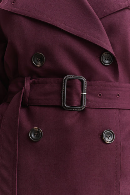 Close-up of a double-breasted trench coat in deep burgundy with a matching waist belt and dark metal buckle.